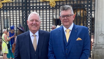 Mike and Kenny stand outside the gates of Buckingham Palace. Both wear dark blue suits with yellow ties.