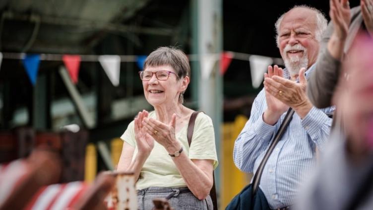 Man and woman are clapping they are standing in front of a string of coloured bunting