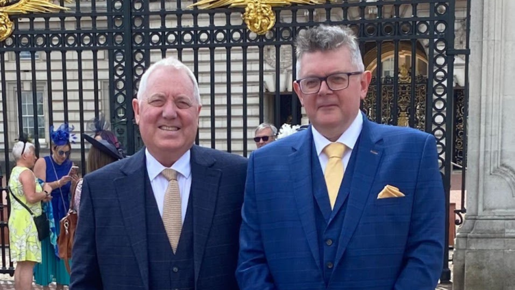 Mike and Kenny stand outside the gates of Buckingham Palace. Both wear dark blue suits with yellow ties.